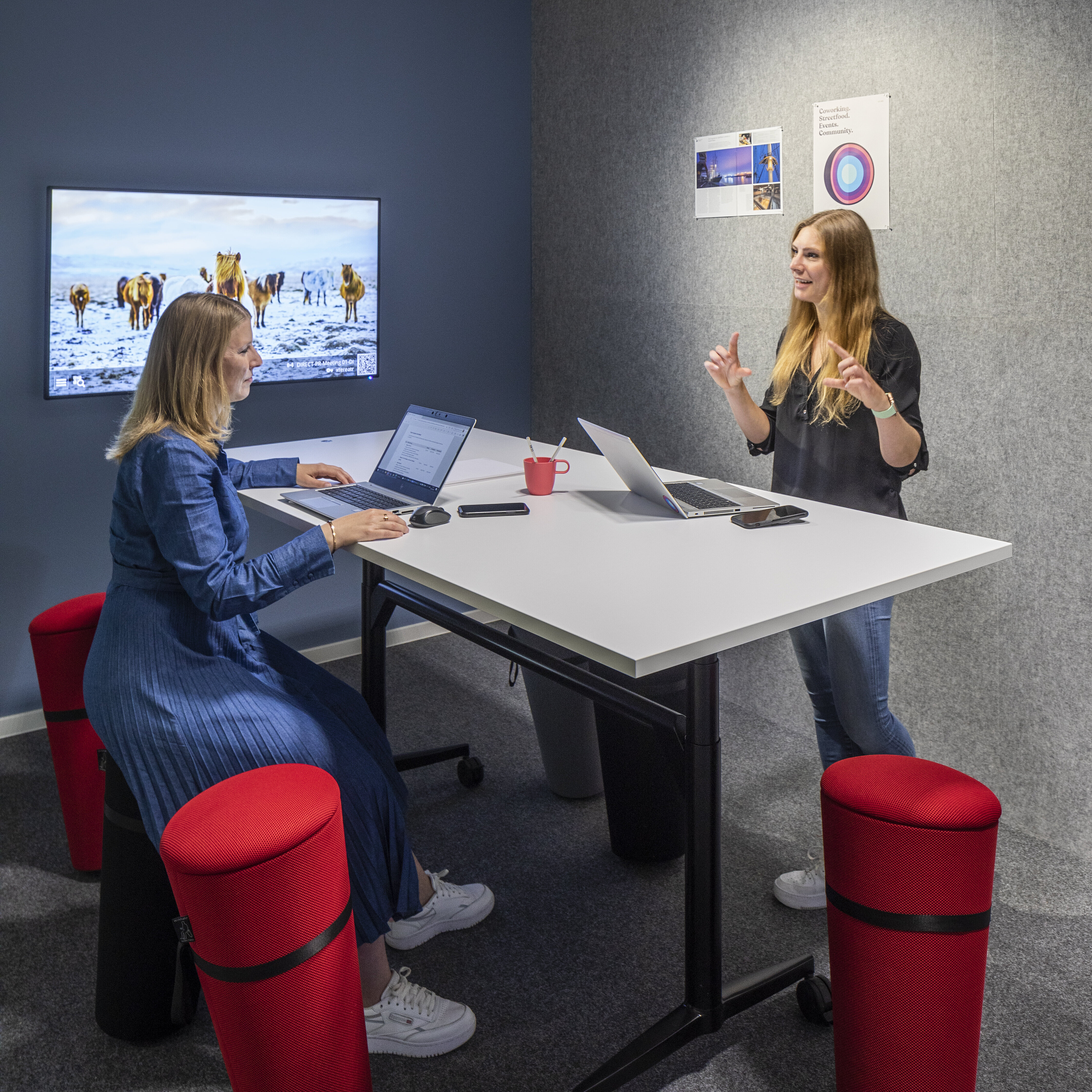 A small meeting room with a standing table, where two people are working intently and watching a presentation on a screen. Clearly visible are several red standing stools, which are used for dynamic work while sitting or leaning. The gray acoustic panels create a quiet, secluded work environment.
