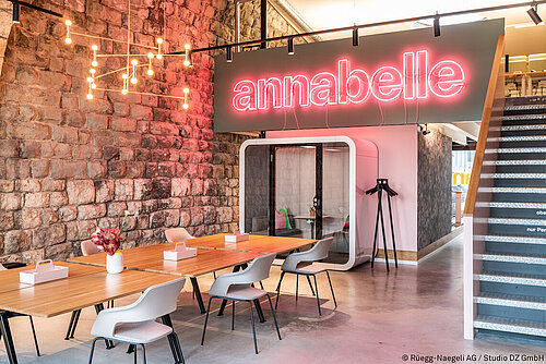 It is an open space with a long table and white Occo chairs on the left. The wall on the left is made of brown brick. On the right, a staircase leads up to the second floor, and on the railing you can see the red-lit lettering “annabelle.”
