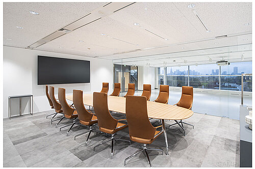 A large conference room is shown here, with a view of the city in the background. The elegant Graph chairs, featuring brown leather and high backrests, lend the room an exclusive, professional atmosphere. The combination of light-colored surfaces, glass, and warm leather creates a high-quality overall look.
