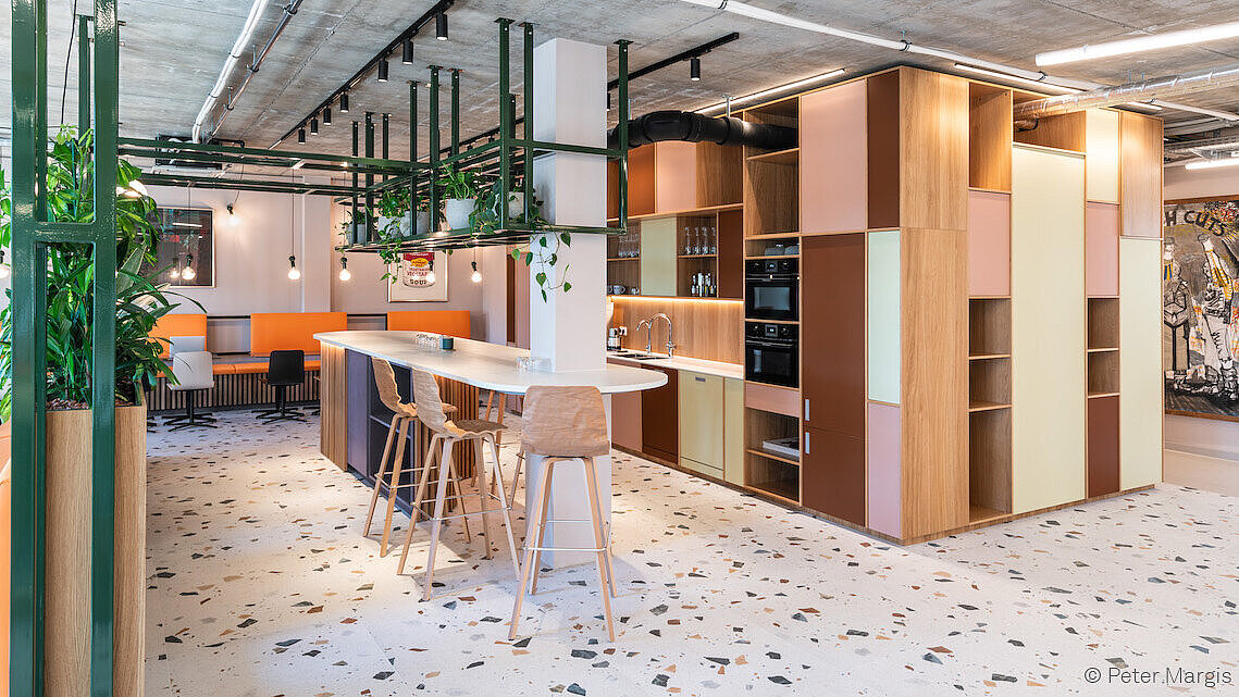 It is an open kitchen with colorful cabinets in shades of red, pink, and white. In front of it is a counter with bar stools. In the background, there is a row of tables along the wall with additional chairs in front of them.