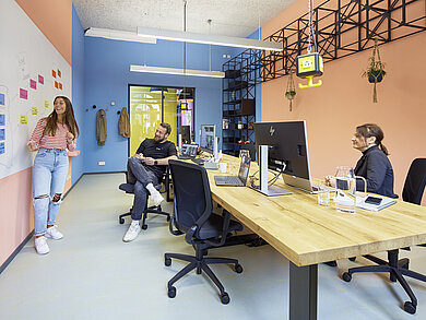 In the room seen in the picture, the walls are orange and blue. In the middle of the room stands a large wooden table, at which two people are sitting on our IN swivel chairs. A third person is standing by the whiteboard on the wall.