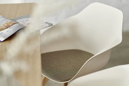 A close-up shows a Yonda chair with a cream-white seat shell, a light brown seat pad and a four-legged wooden base, standing next to a table with a light-colored tabletop. An open magazine lies on the table.