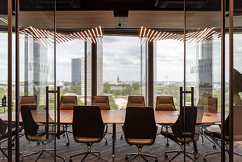 The conference room features a long table set with Graph chairs upholstered in brown leather and featuring high backrests. The large windows create an elegant, light-filled atmosphere with a view of the city. The striking ceiling lighting adds a modern, sophisticated touch to the room.