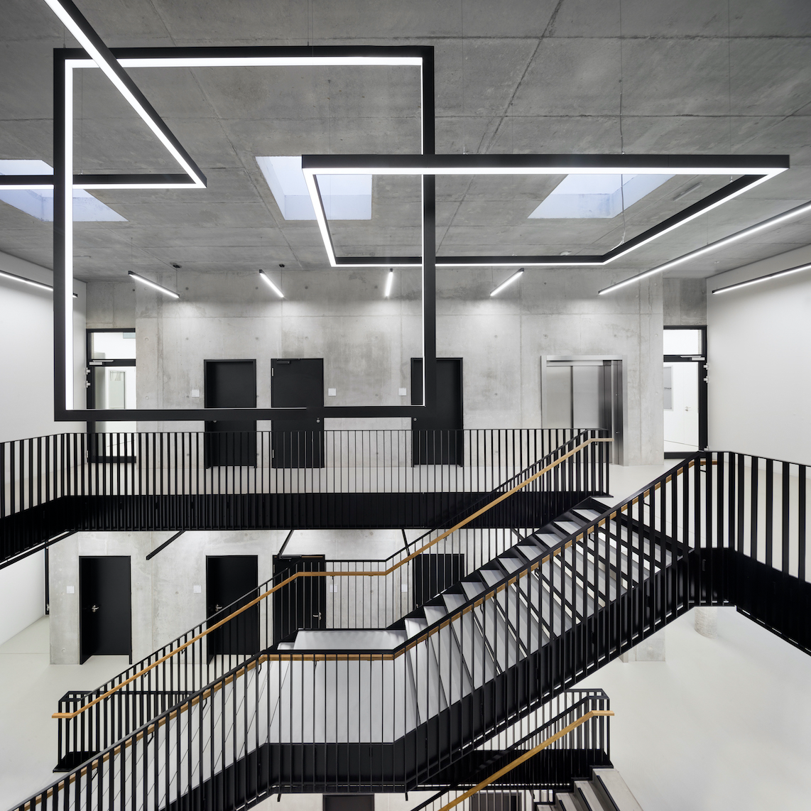 The photo shows a large, modern stairwell with clean lines and a striking LED light installation on the ceiling. Exposed concrete, black railings, and light-colored floors create an industrial, minimalist look.