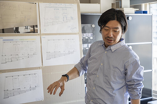 The photo shows a man in a work or design room standing in front of several architectural or floor plan drawings hanging on the wall, explaining something. The scene conveys a creative, planning-oriented work process.