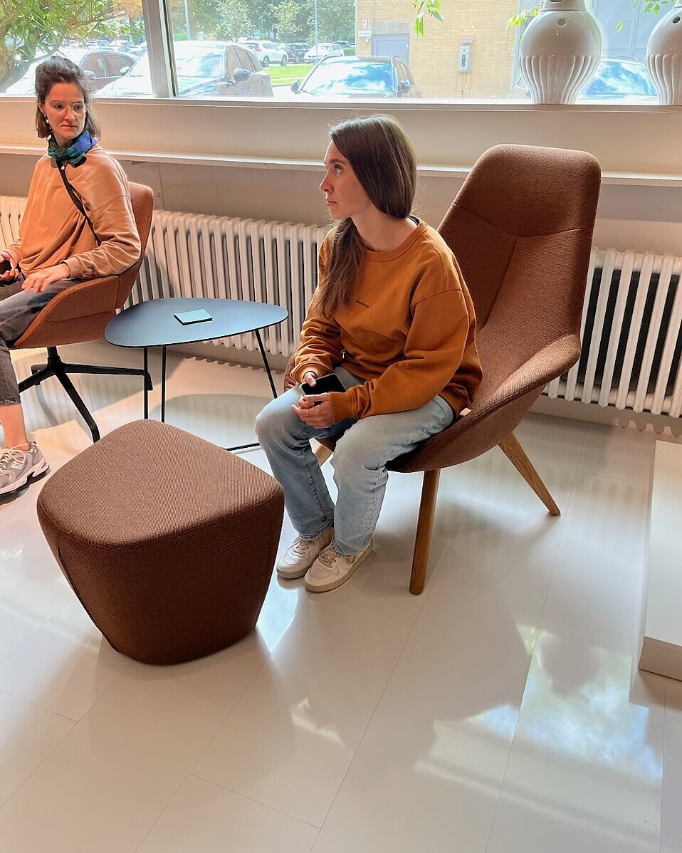 The two women are sitting in comfortable Yonda lounge chairs in a warm brown shade. The organic shapes and wooden frame create a cozy, relaxed atmosphere. The scene depicts a casual conversation in a modern lounge area.