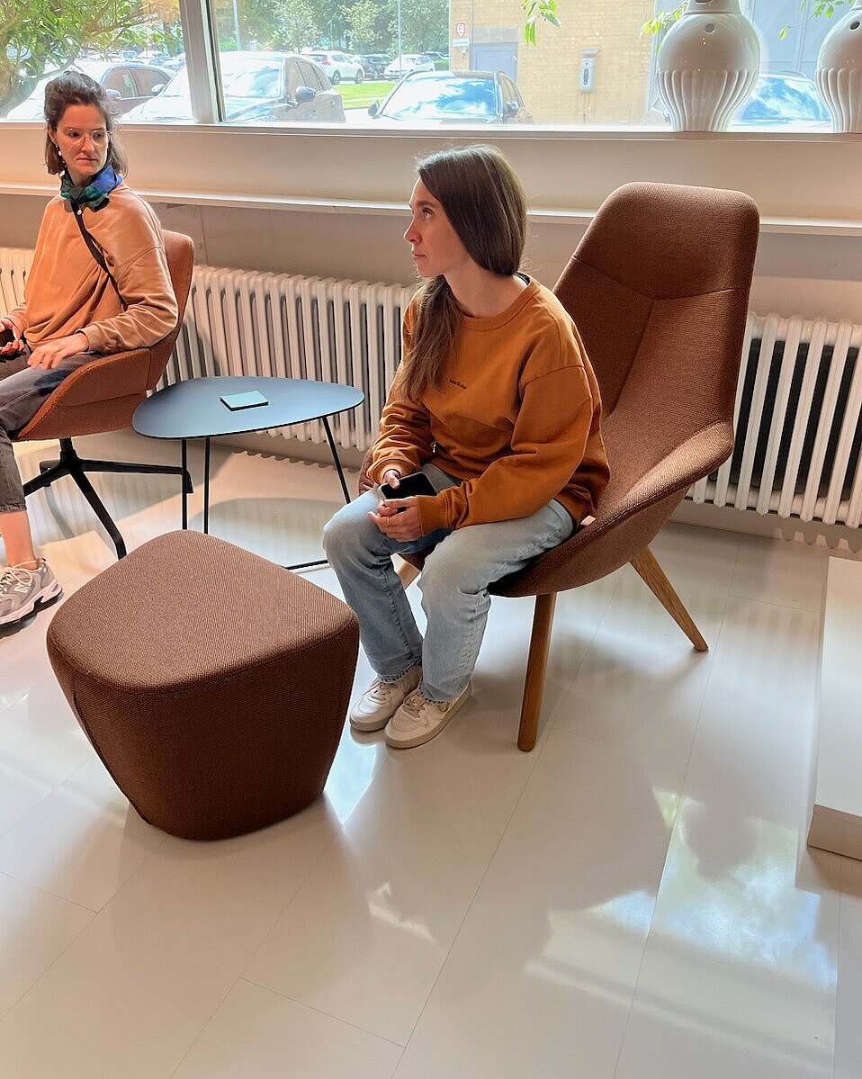 The two women are sitting in comfortable Yonda lounge chairs in a warm brown shade. The organic shapes and wooden frame create a cozy, relaxed atmosphere. The scene depicts a casual conversation in a modern lounge area.