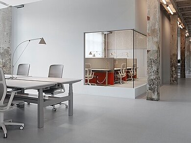 The open workspace combines industrial charm with modern office design. Several AT Mesh units in light gray are on display, blending harmoniously into the minimalist setting. The furniture’s clean lines complement the calm, functional design of the space. 