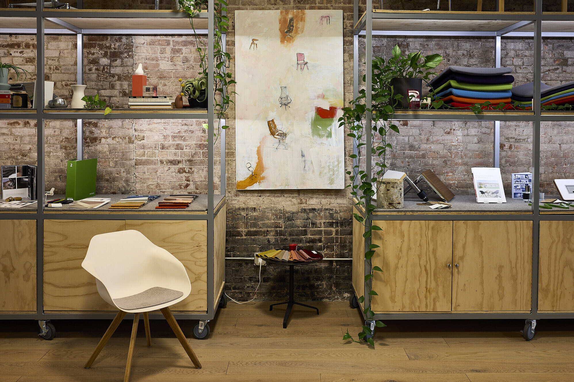 A Yonda chair with a white seat shell, a gray felt pad, and a four-legged wooden base stands in front of two shelves filled with various Wilkhahn samples. These include fabrics, sample books, upholstery, and table surfaces. The room is decorated in an industrial style.