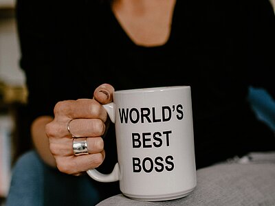A close-up of a woman sitting in a chair. The focus is on the white mug in her hand with the phrase “World's best boss”