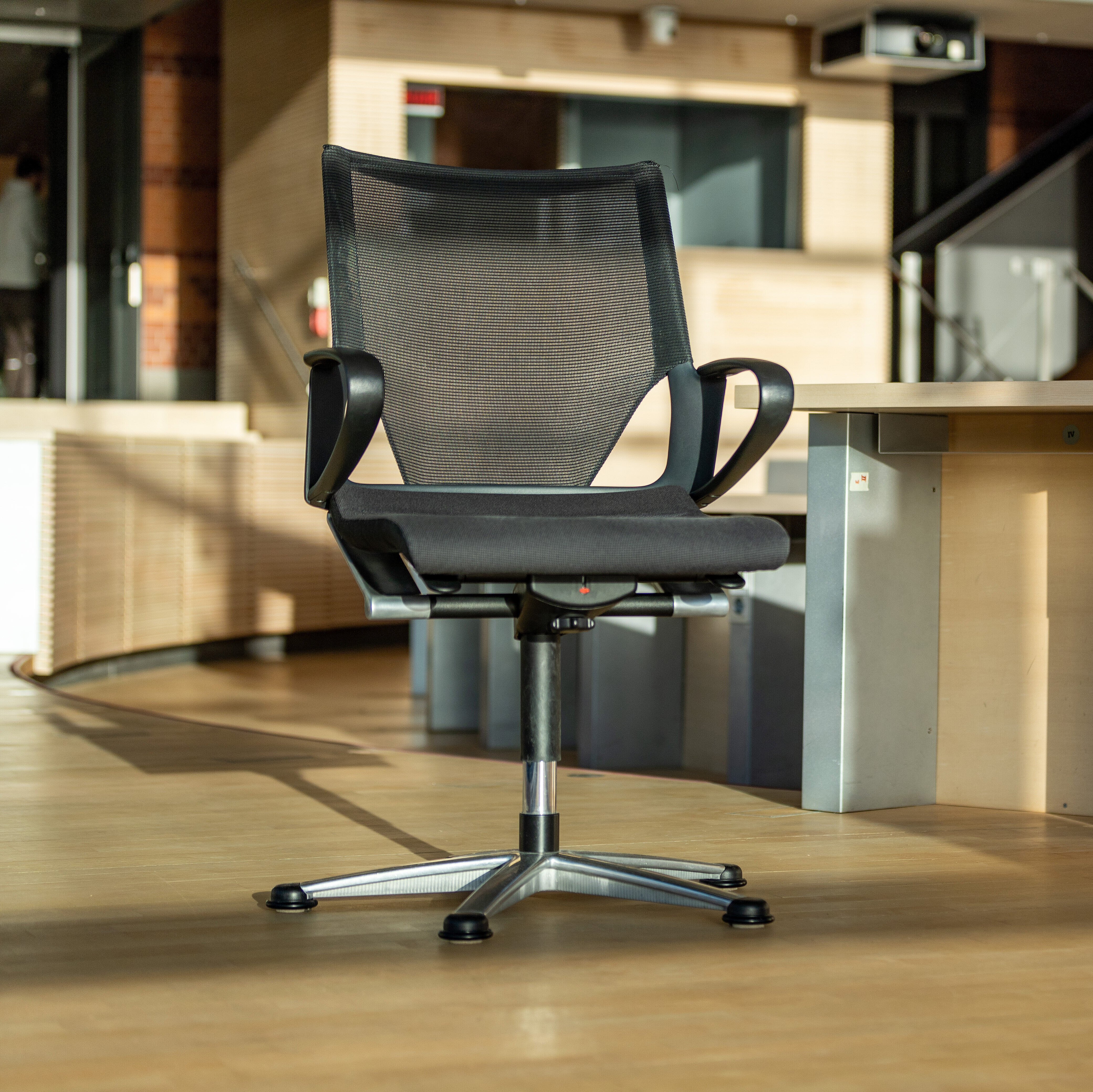 A black Modus swivel chair stands in a room where one would otherwise see only rows of wooden tables.
