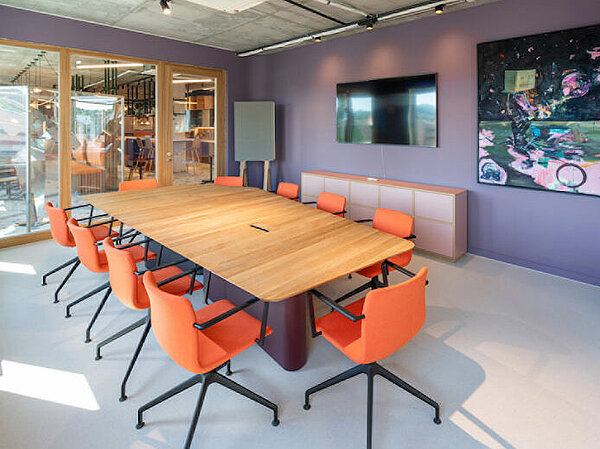 The photo shows the same conference room from two different angles: one taken from the right side and one from the left. The walls are painted purple, and in the center stands a large wooden table with Pep chairs around it, which have black frames and orange upholstery.