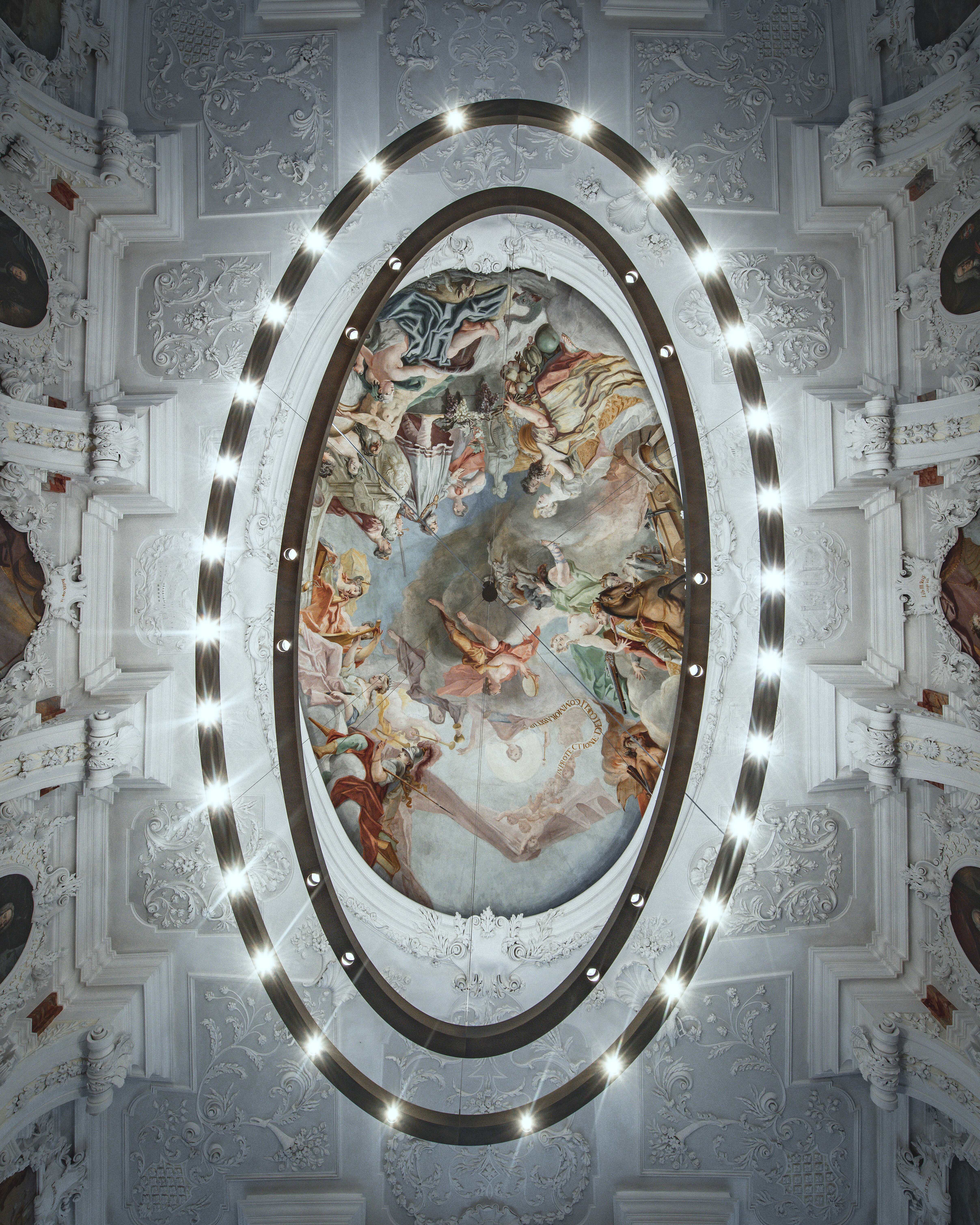 A close-up of a circular ring lamp. In the center is an elaborate wall painting showing various gods.