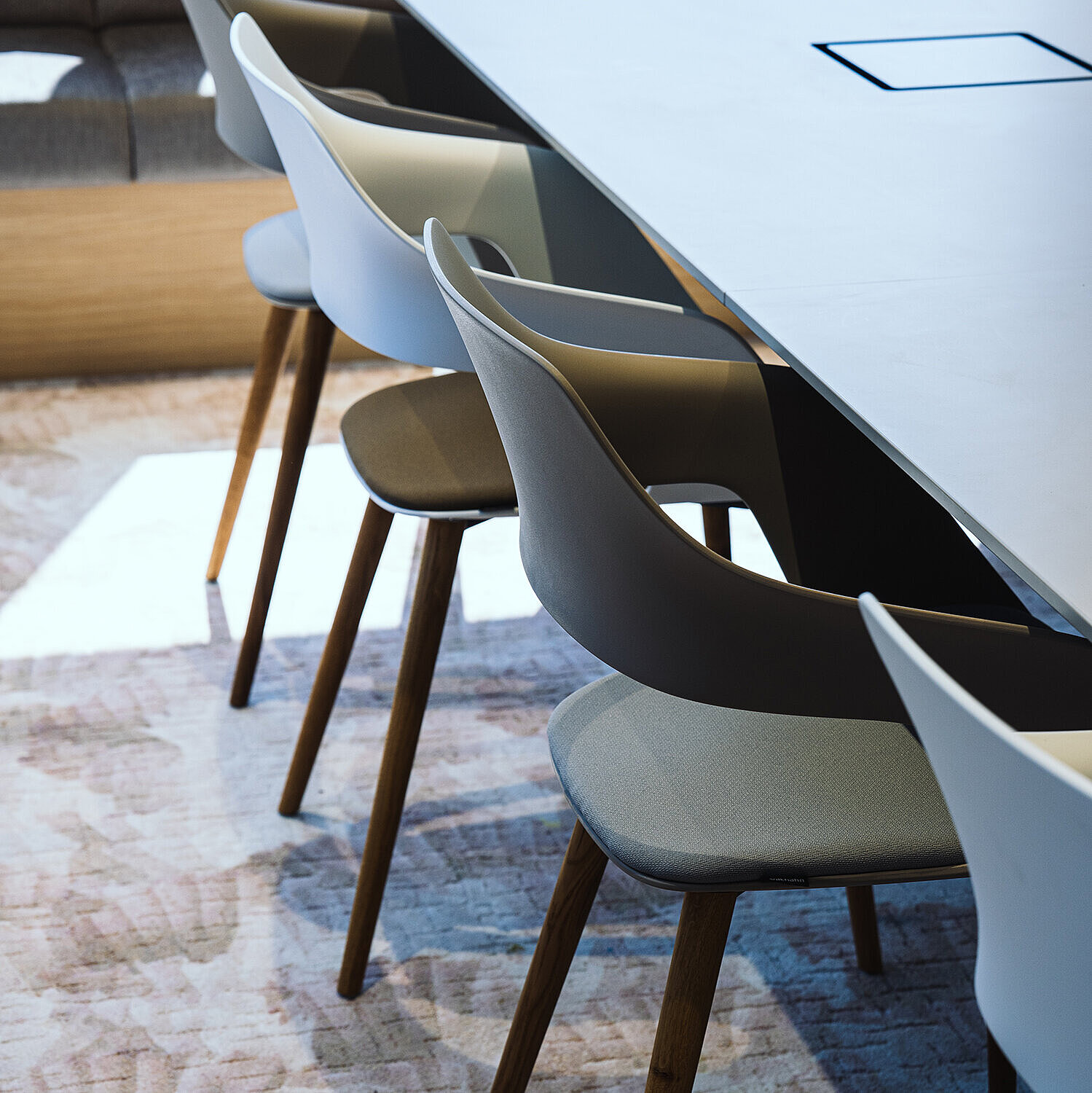 Occo chairs with white or gray seat shells and seat pads in shades of green are arranged around a white table. Die Stühle haben ein Vierfußgestell aus Holz.