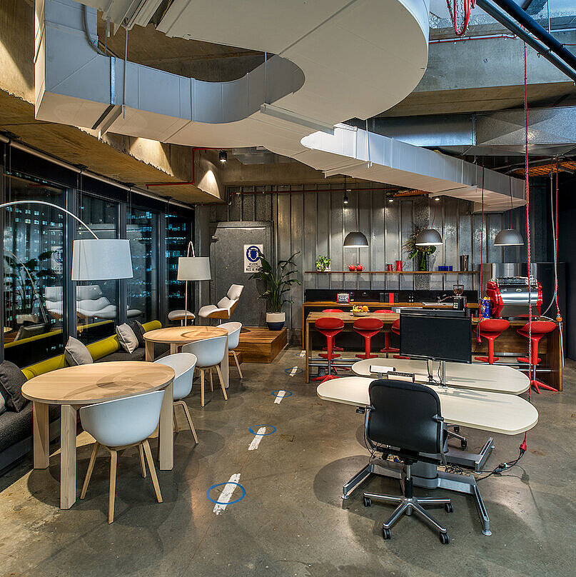 The photo shows a modern, open-concept office with various work and seating areas. In the foreground is a workstation featuring a black FS swivel chair. The industrial interior design is complemented by colorful accents and comfortable furniture.