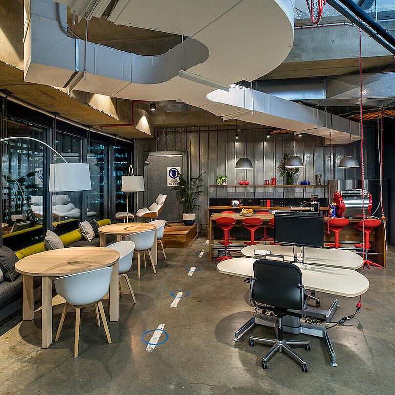 The photo shows a modern, open-concept office with various work and seating areas. In the foreground is a workstation featuring a black FS swivel chair. The industrial interior design is complemented by colorful accents and comfortable furniture.