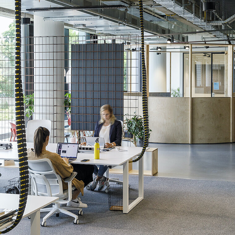 An open-plan office with high ceilings, exposed building services, and a modern, airy working atmosphere. Two people are working at a light-colored desk; in the foreground is a white AT Mesh. Plants and modular room dividers provide structure within the open space.