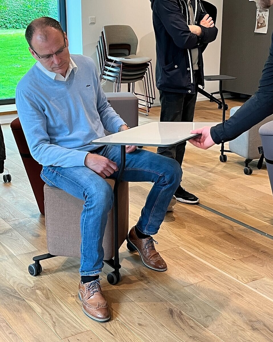 A man is testing a rolling stool with a built-in swivel tray. The setup demonstrates its versatile use for workshops and agile work formats. The scene emphasizes mobility, experimentation, and ergonomic adjustability.