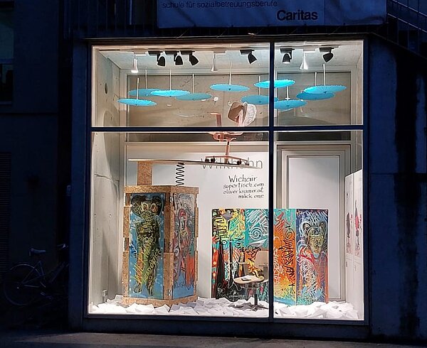 This is an exterior view of a building with a large storefront window. The window shows many colorful graffiti images and our WiChair. The seat and backrest are made of light-colored wood.