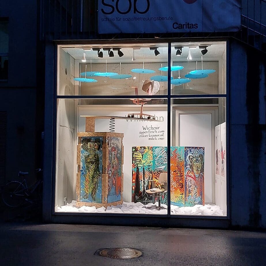 This is an exterior view of a building with a large storefront window. The window shows many colorful graffiti images and our WiChair. The seat and backrest are made of light-colored wood.