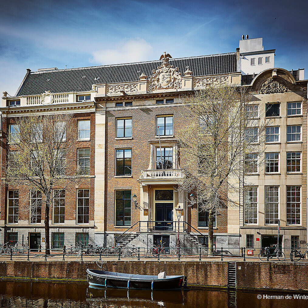This is an exterior view of a building with a brick facade. There are many bicycles parked at the building's entrance. In front of it, you can see a river with a boat on it.