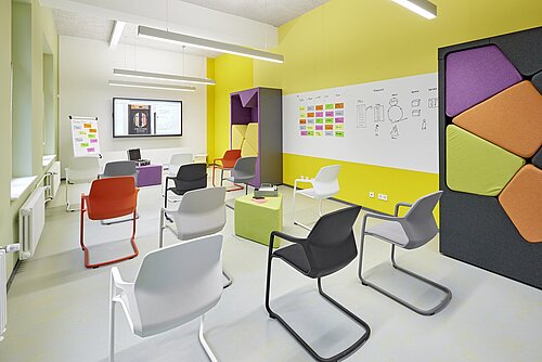In a very colorful room with yellow and green walls, there are many Metrik chairs in various colors. For example, in red, white, beige, black, and gray.
