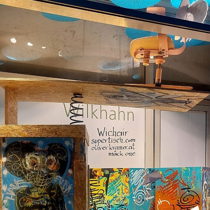 It is a picture of a very large vitrine containing with various colorful graffiti paintings and with our WiChair without armrests in front of it. The seat and backrest are made of light-colored wood.
