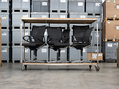 Several black swivel chairs are on the lower level of a double-decker carriage. In the background, gray boxes can be seen in a warehouse.
