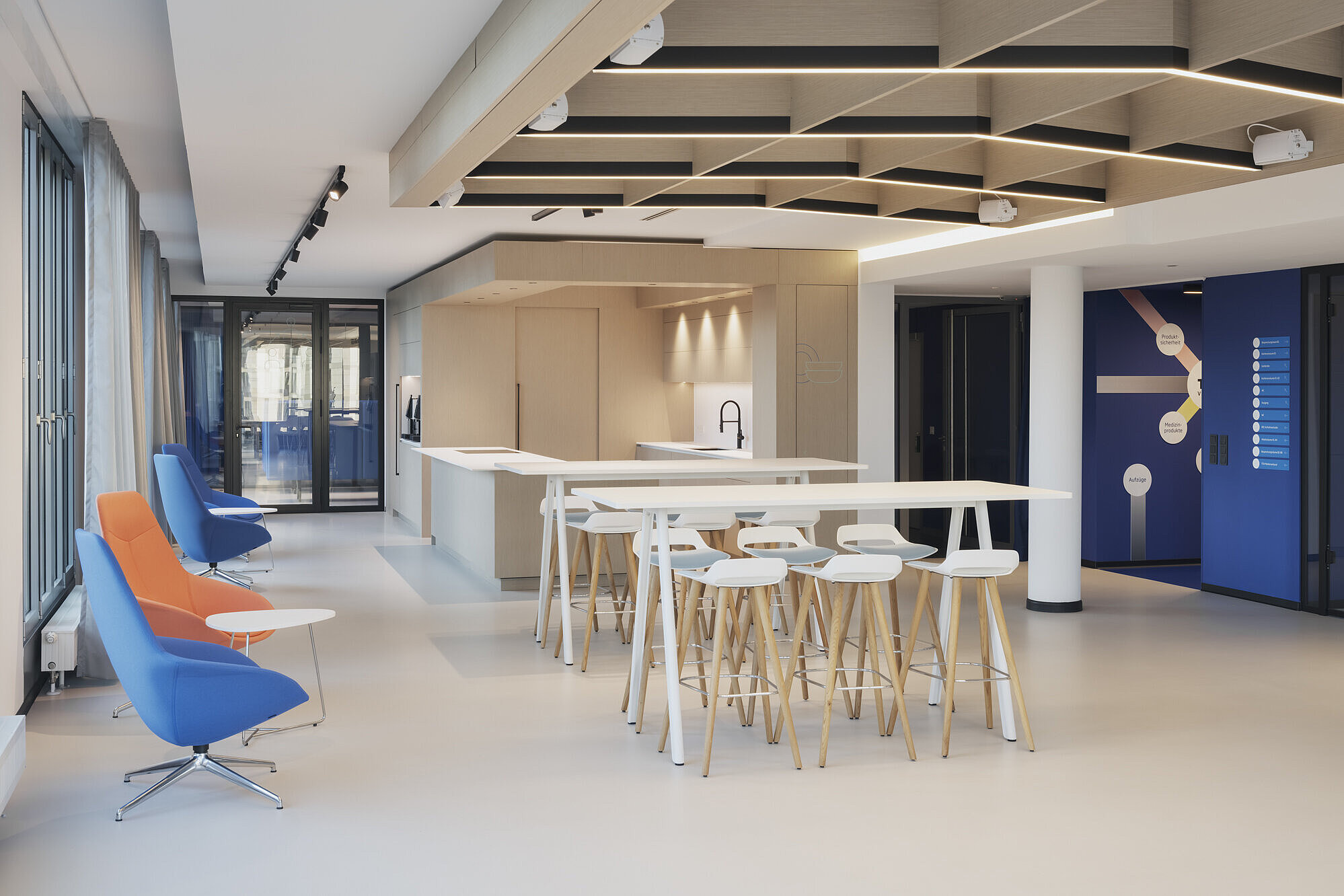 The open lounge and gathering area features several colorful Yonda lounge chairs upholstered in blue and orange. The tall tables and Occo bar chairs complement the modern, inviting space. The light-colored architecture enhances the open, welcoming atmosphere.