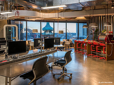 The photo shows a modern office with large windows offering a view of the city. In the foreground are several workstations equipped with black FS swivel chairs. The exposed wiring and ventilation ducts on the ceiling give the room an industrial look.
