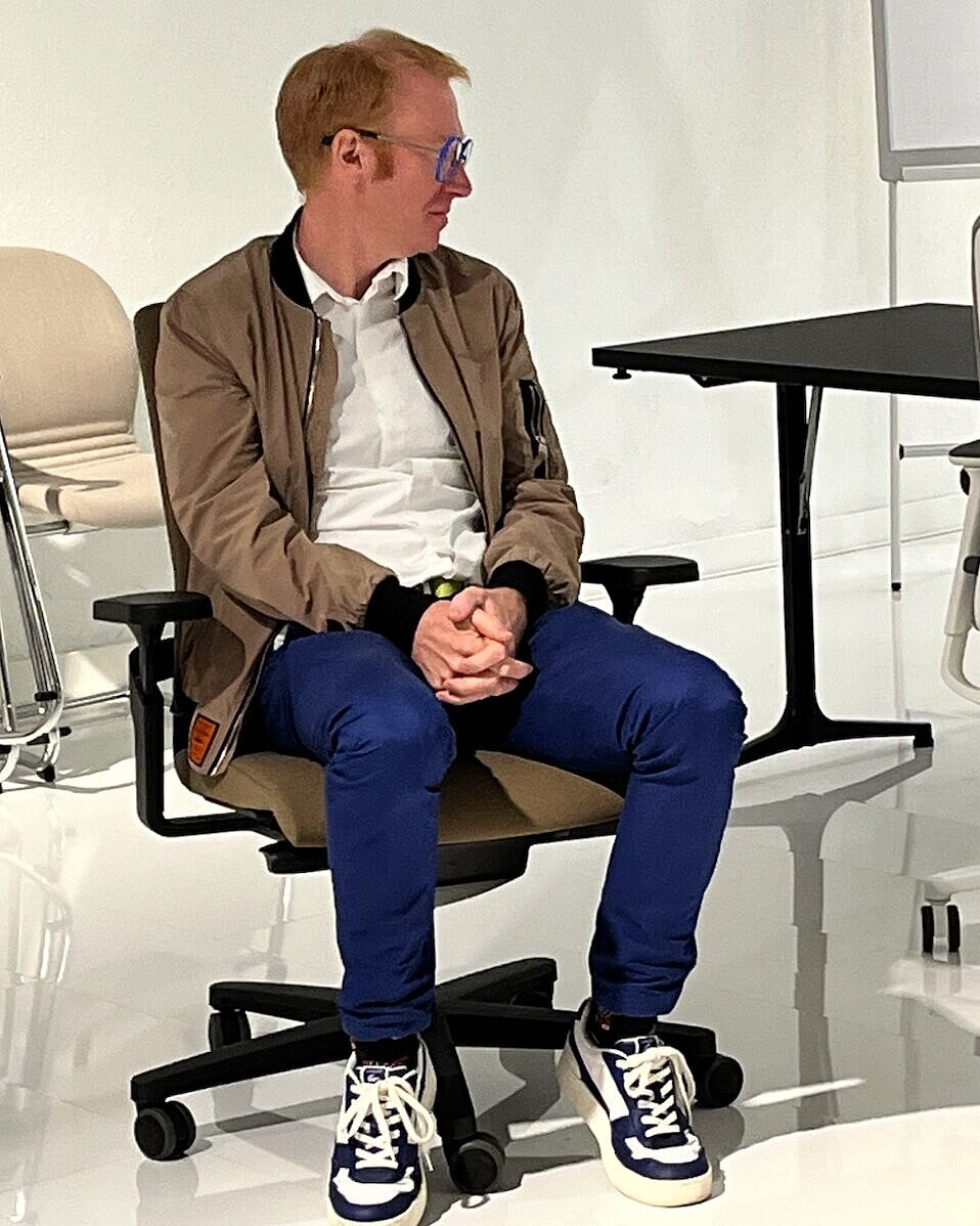 The man is sitting on an AT chair with a black frame and brown upholstery. The neutral setting highlights the chair and its comfort features. The scene looks as though he is testing or inspecting the chair's comfort.