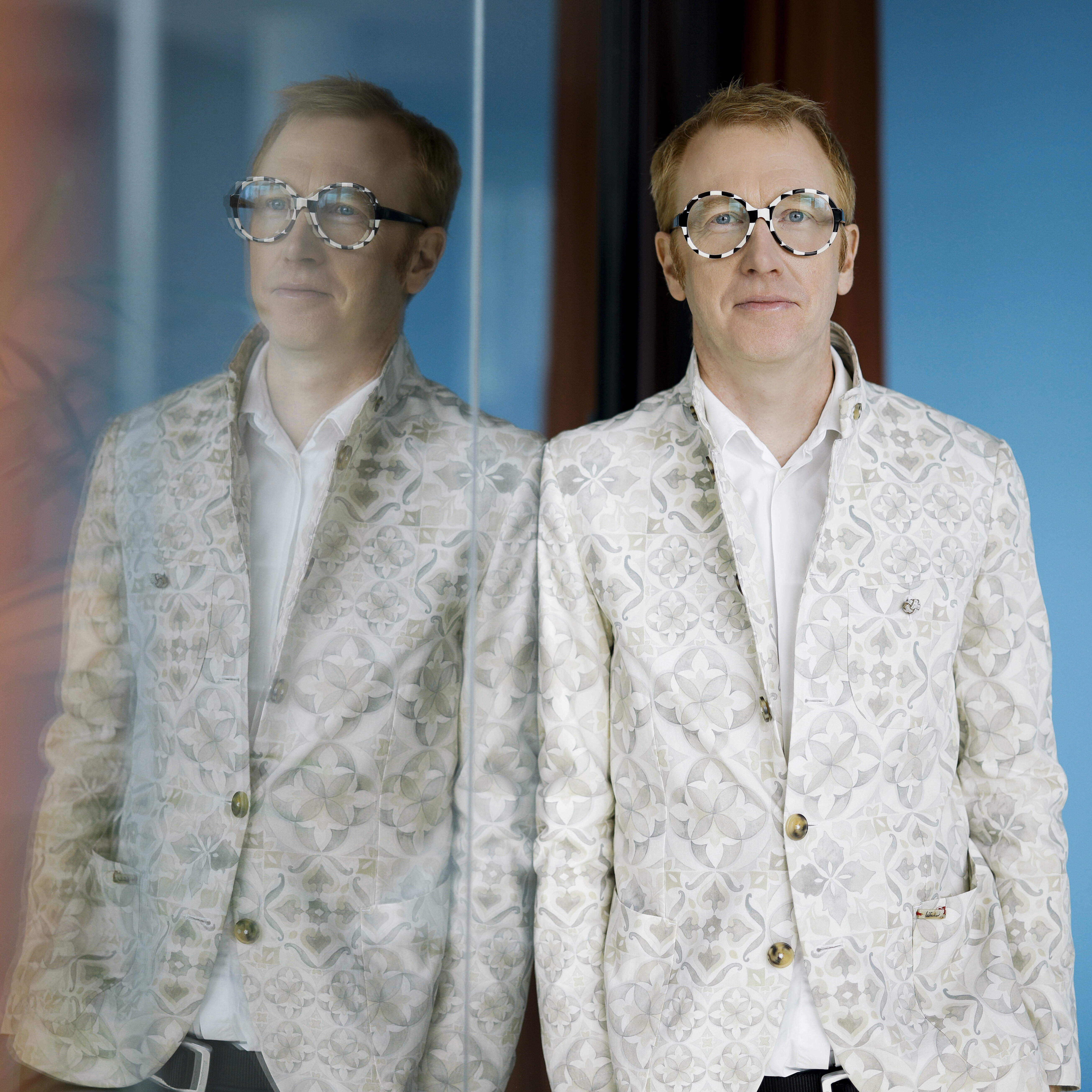 The portrait depicts a man in a creative business outfit, set against a colorful, modern office backdrop. The image’s impact comes from his personality and the reflection in the glass.