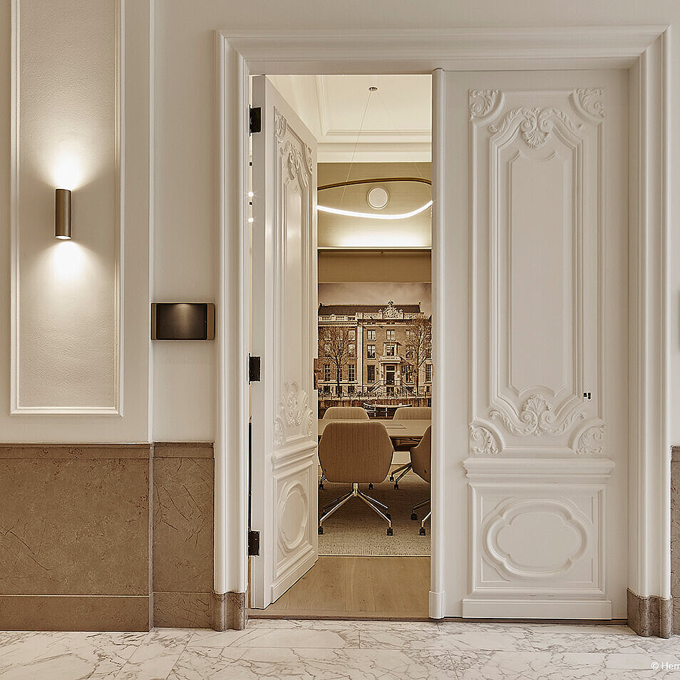 It opens onto a modern meeting room with elegant conference chairs through a richly decorated, classic double door. The warm, upholstered swivel chairs are only partially visible.