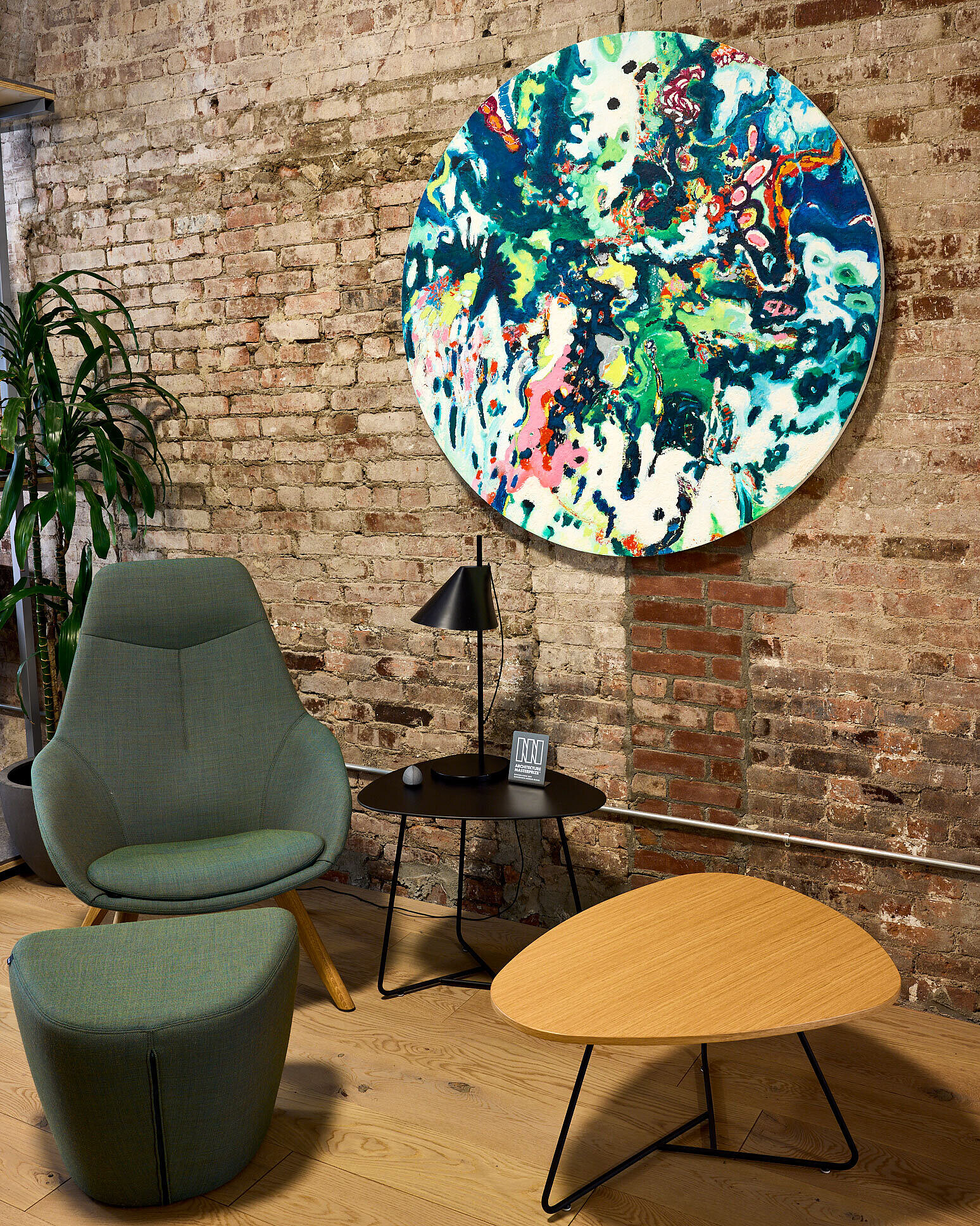 A green, full-upholstered Yonda lounge chair with a matching upholstered footstool stands in front of a brown brick wall. Next to it are two Yonda lounge tables with black cantilever bases. One has a wooden tabletop, the other a black one.