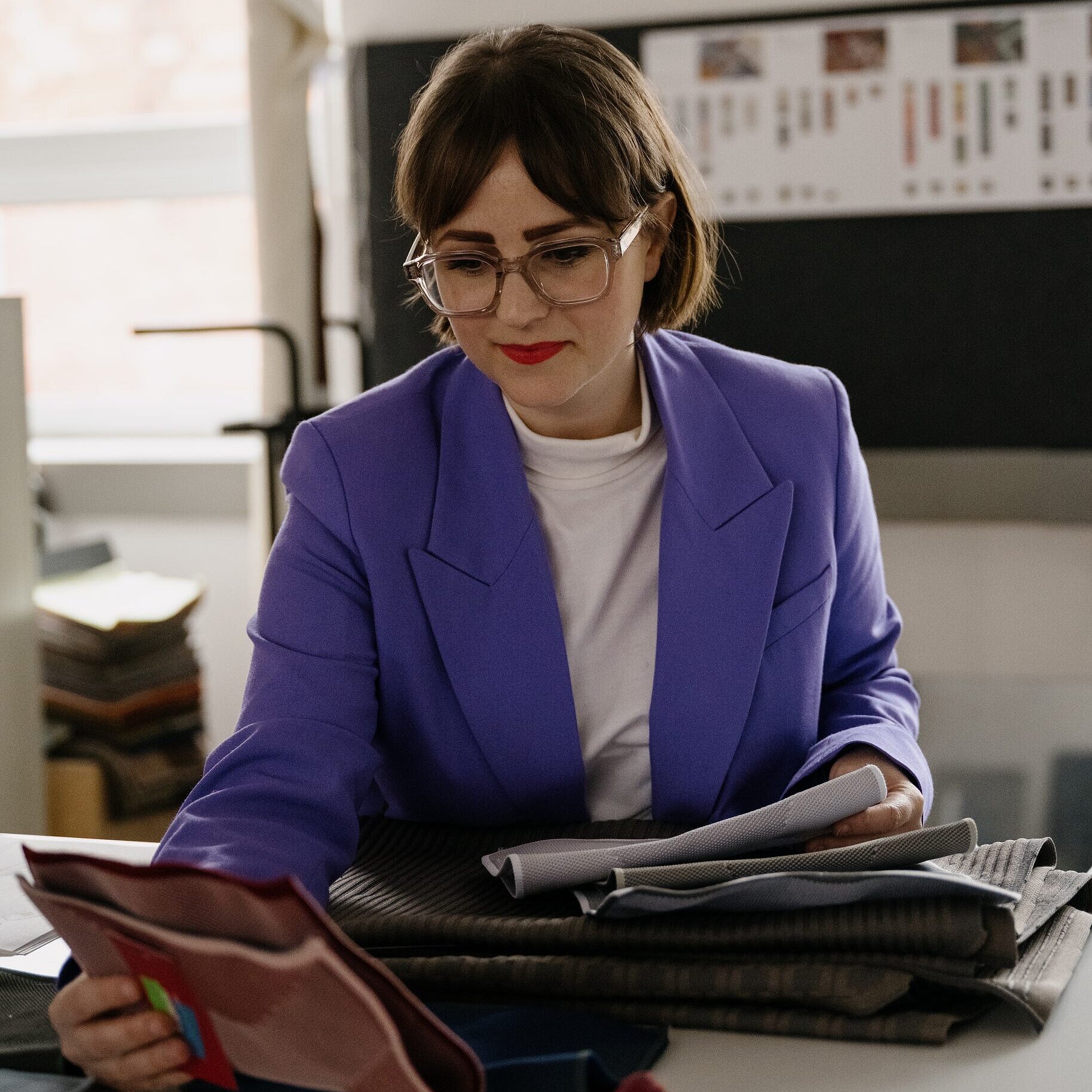 Ms. Schnizler, wearing a white T-shirt and a purple blazer, is standing at a table looking at various fabric samples.