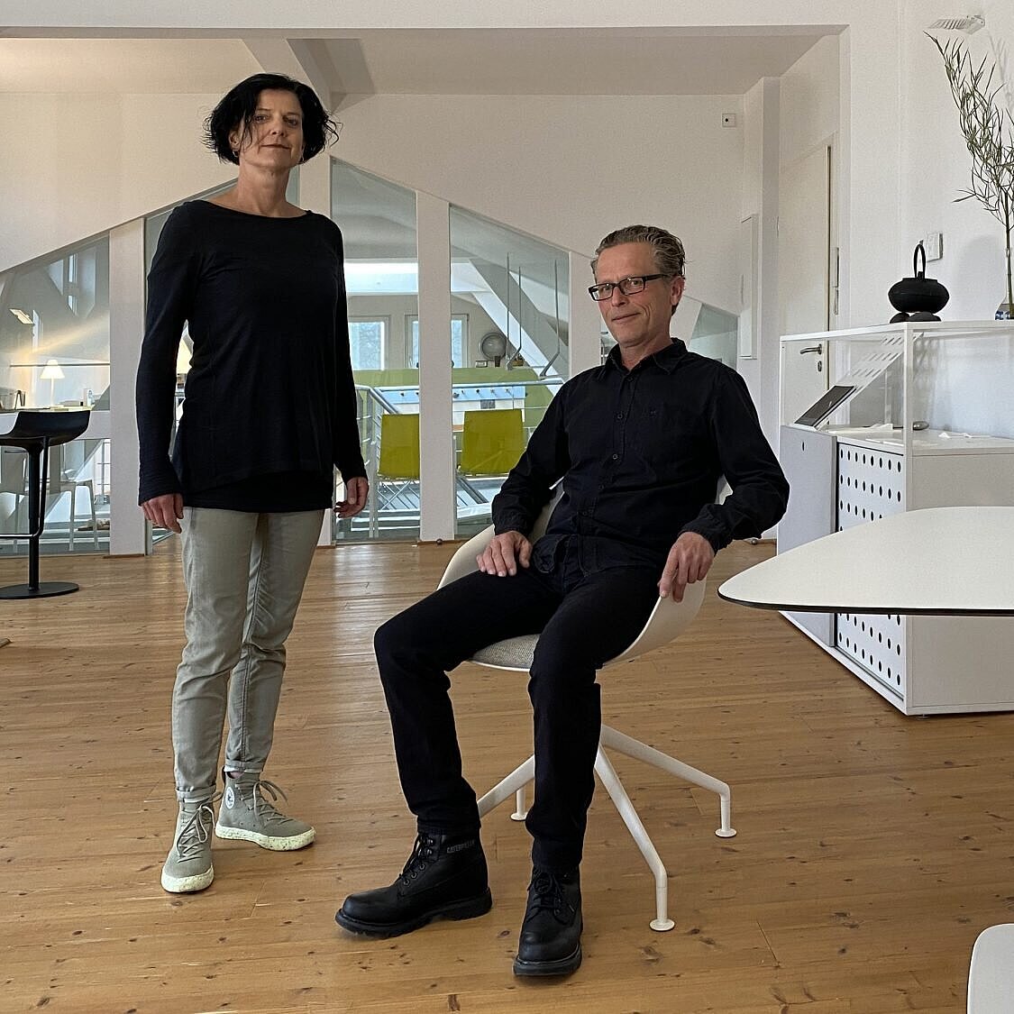The two people are in a bright, modern office or studio space. The man is sitting on a Yonda, recognizable by its distinctive shell-shaped seat and delicate base. There are also Yonda design models on the table in the foreground.