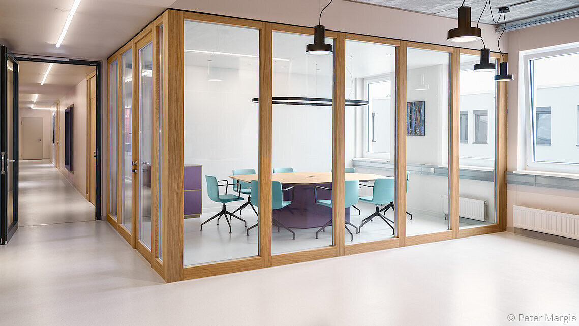 A conference room was photographed from the outside. The walls consist of large windows. Inside the conference room, there is a wooden table and Pep chairs with black frames and turquoise upholstery.