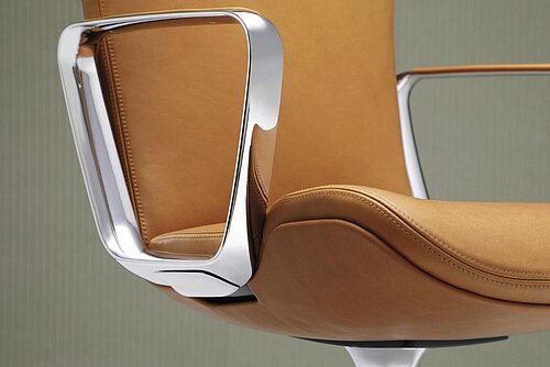 This close-up shows the elegant armrest of a Wilkhahn Intra conference chair in polished aluminum. The high-quality leather upholstery in a warm cognac tone emphasizes the chair’s luxurious appearance and precise craftsmanship. The focus is clearly on material quality and design craftsmanship. 