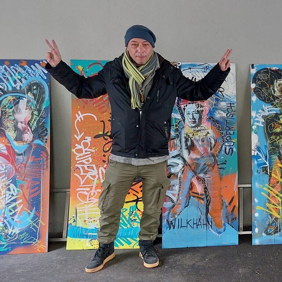 A man wearing a dark blue jacket and a cap is standing in front of several graffiti-style paintings, some of which feature the Wilkhahn logo. The paintings are very colorful, with bright, eye-catching colors.