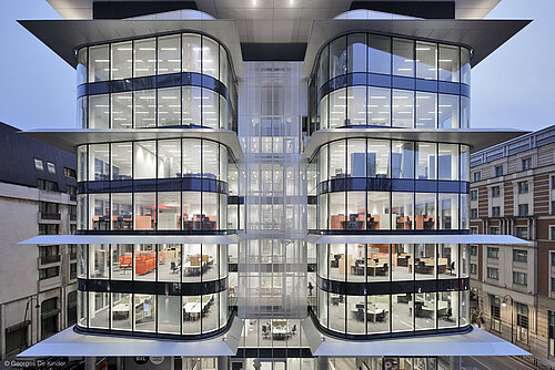 The exterior of a very modern building. Through the many windows, you can see the individual offices and a few AT swivel chairs.