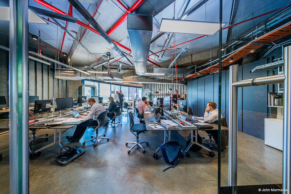 In a large office, several people can be seen going about their work. There are a few black FS swivel chairs in the room. In the background is a large window. Up near the overhead, you can see pipes and cables.