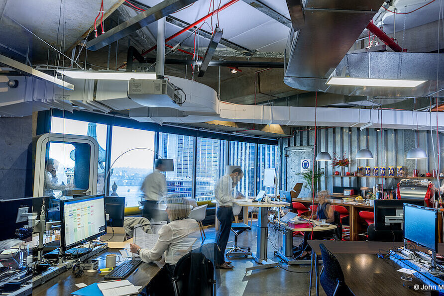In a large office, several people can be seen going about their work. There are a few black FS swivel chairs in the room. In the background is a large window. Up near the overhead, you can see pipes and cables.