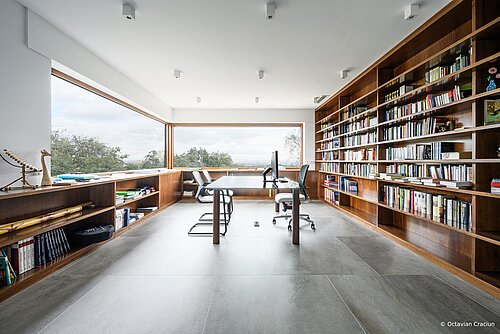 The photo shows a bright home office with large panoramic windows offering a sweeping view of the surrounding greenery. Two gray Metrik cantilever chairs are placed around the glass table. Opposite them stands a gray-black IN swivel chair.