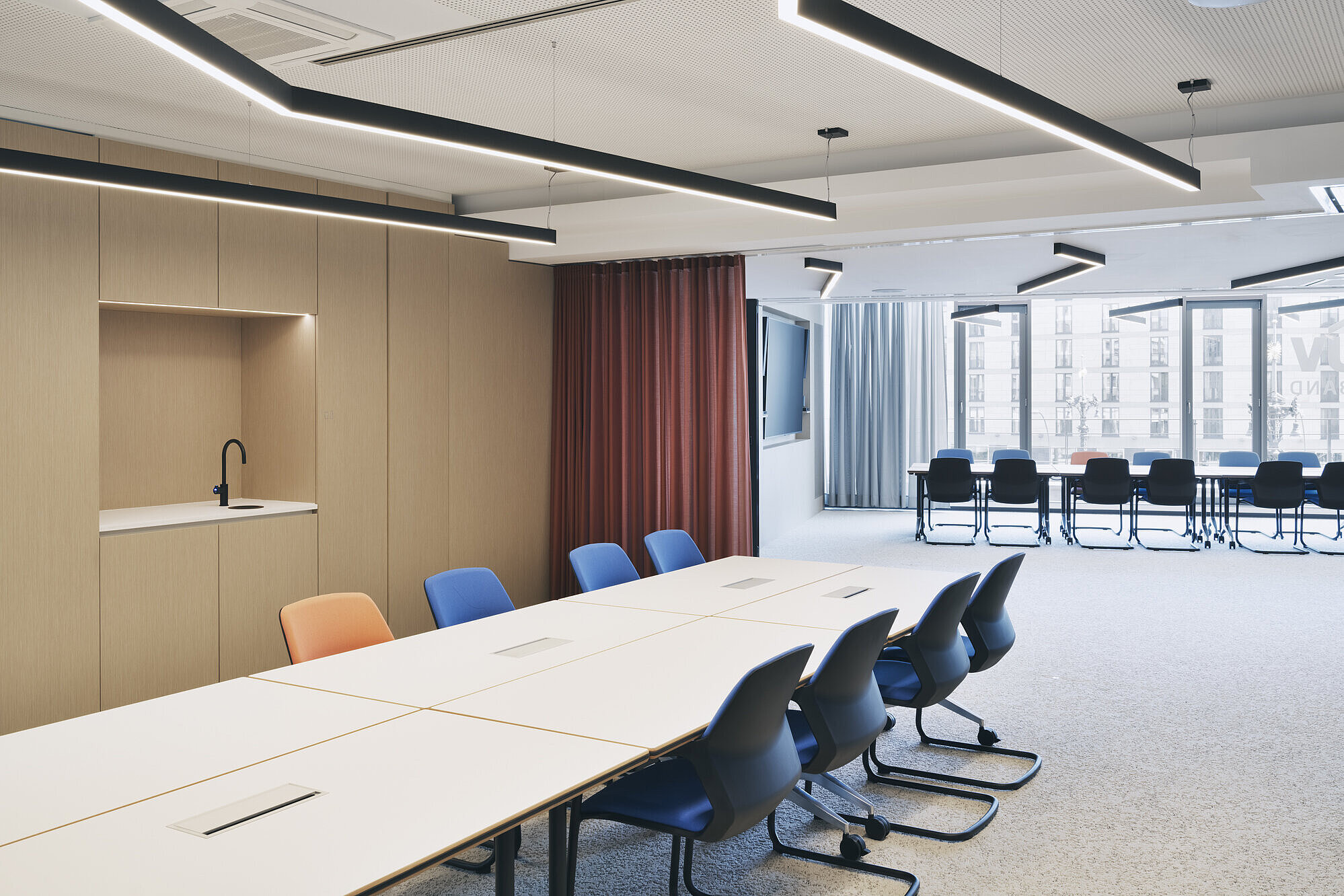 The conference room features a row of Metrik cantilever chairs in blue and orange, which stand out for their organic shape and comfortable upholstery. The flexible table arrangements and warm materials create a modern, professional meeting environment. Large windows provide plenty of natural light.