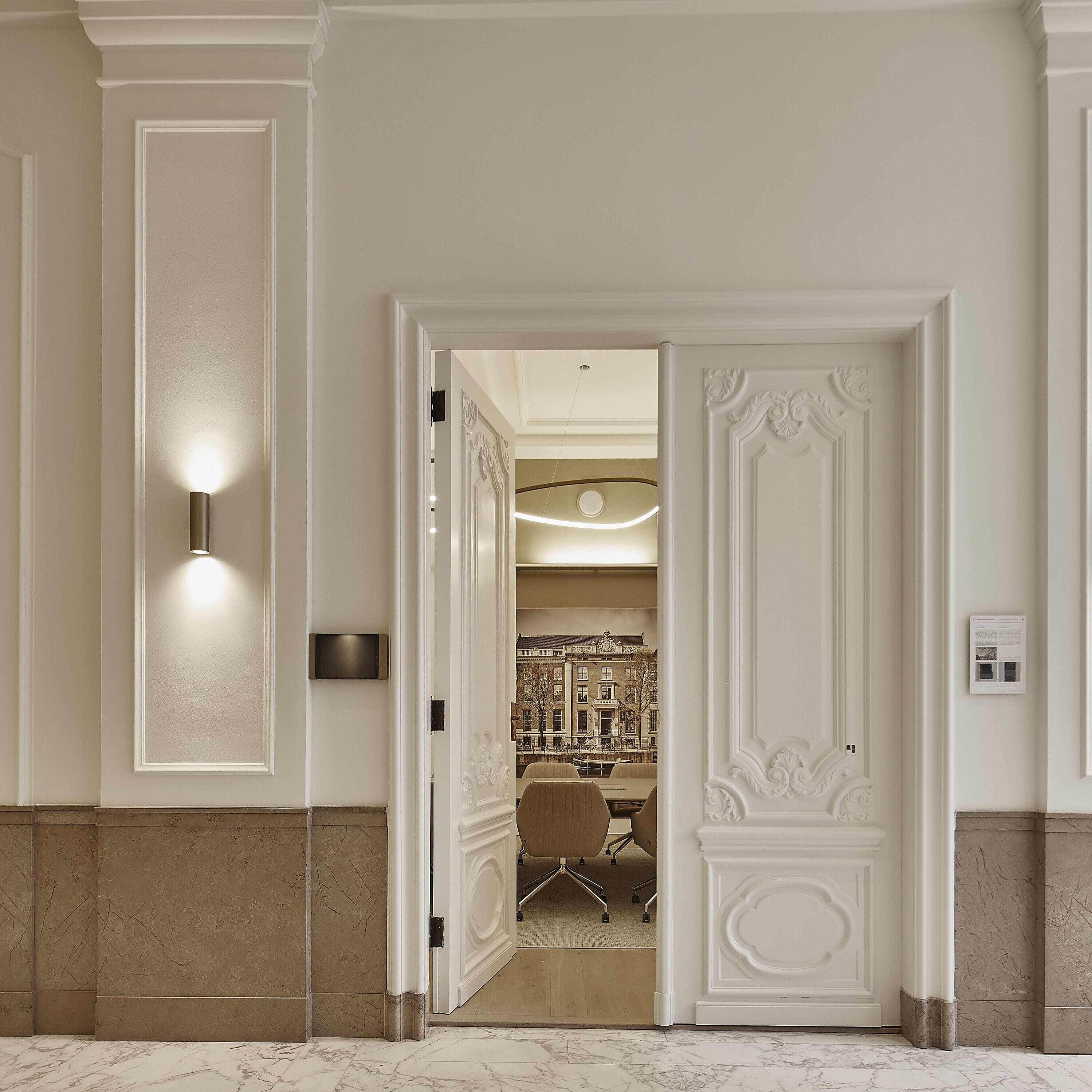 It opens onto a modern meeting room with elegant conference chairs through a richly decorated, classic double door. The warm, upholstered swivel chairs are only partially visible.