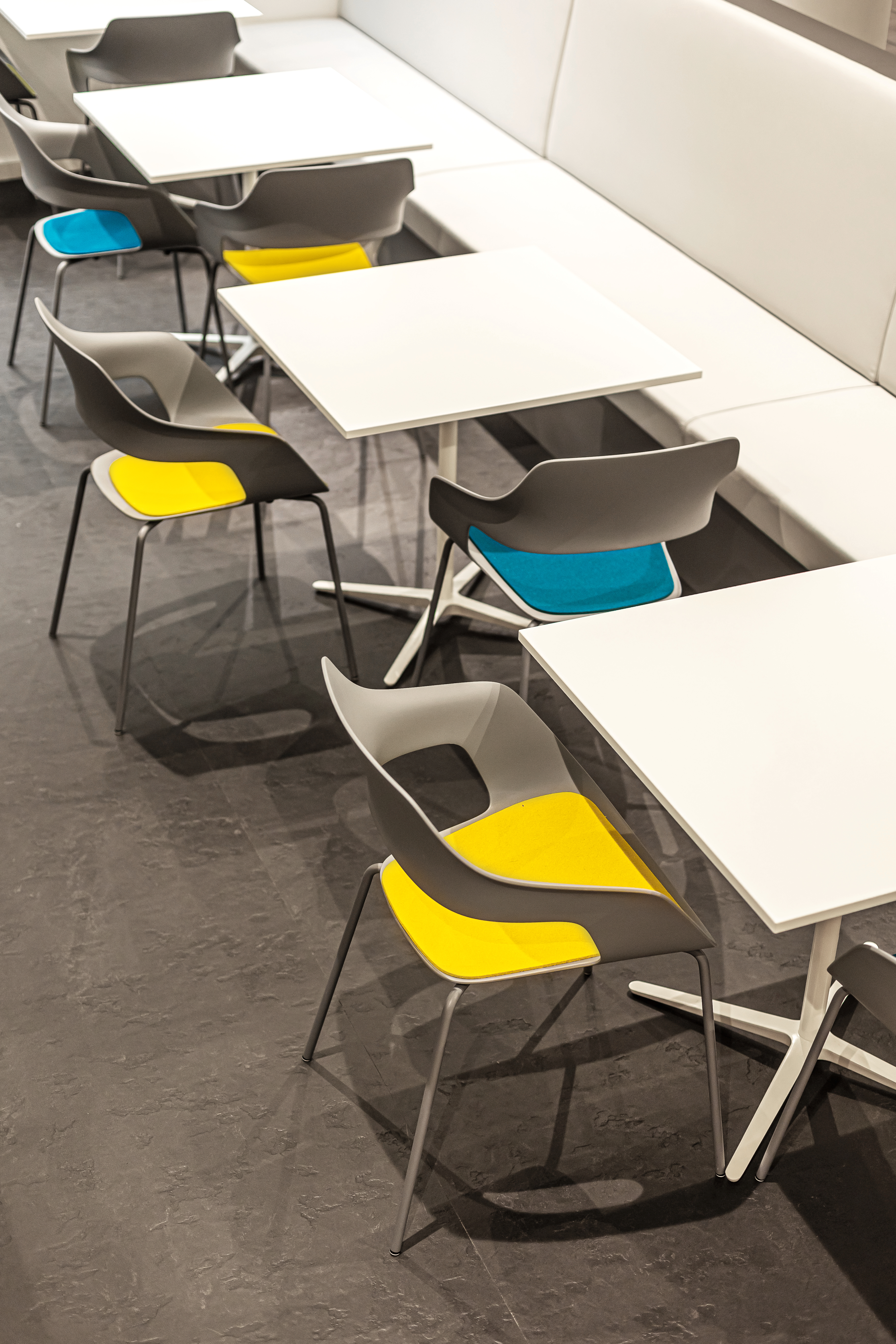 This is a view of a section of the cafeteria where Occo chairs with yellow and blue seat pads are placed at the tables, which can be seen in front of white benche seats.