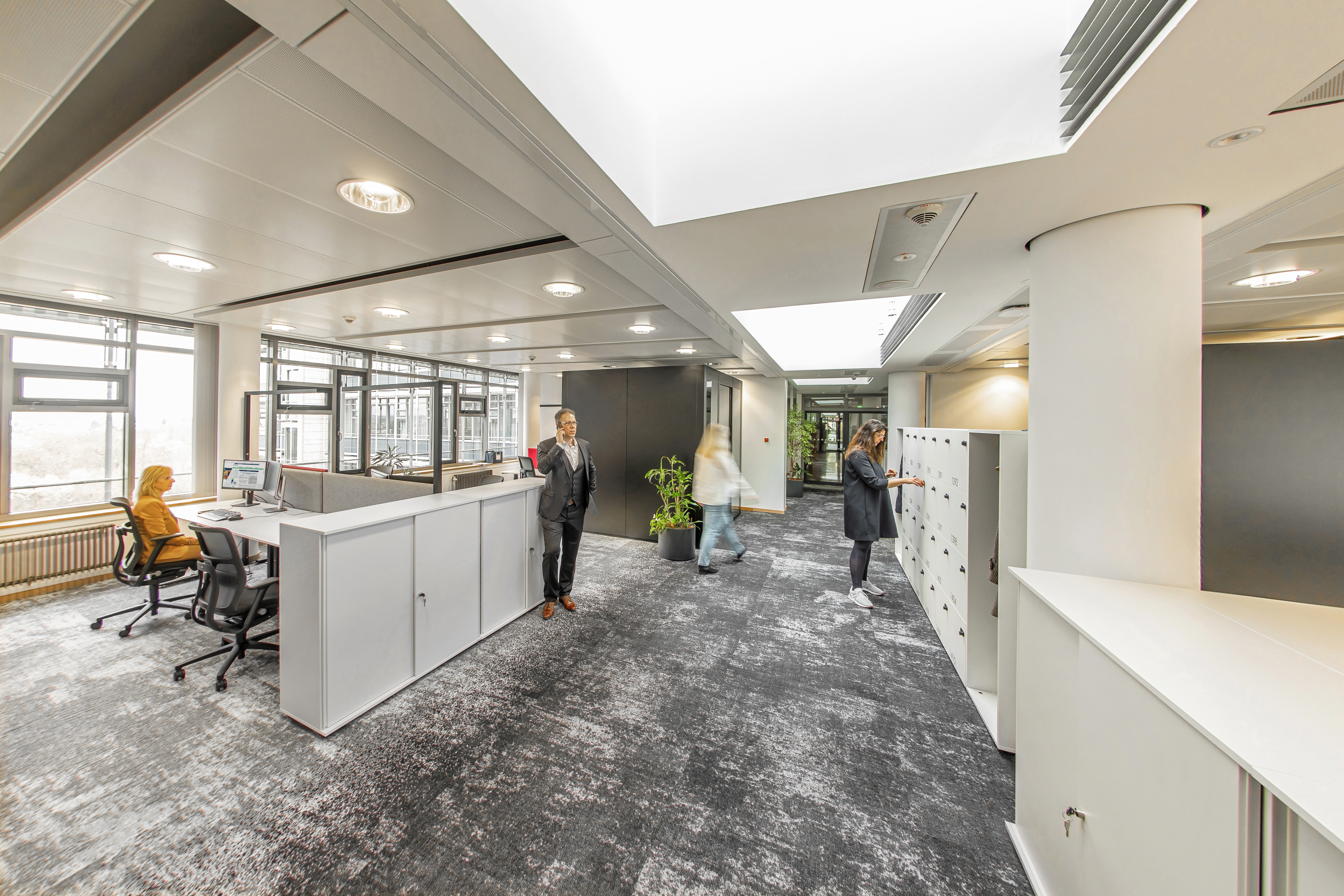An office with white shelves on the right and a table island on the left. Black AT swivel chairs are placed around the table island.