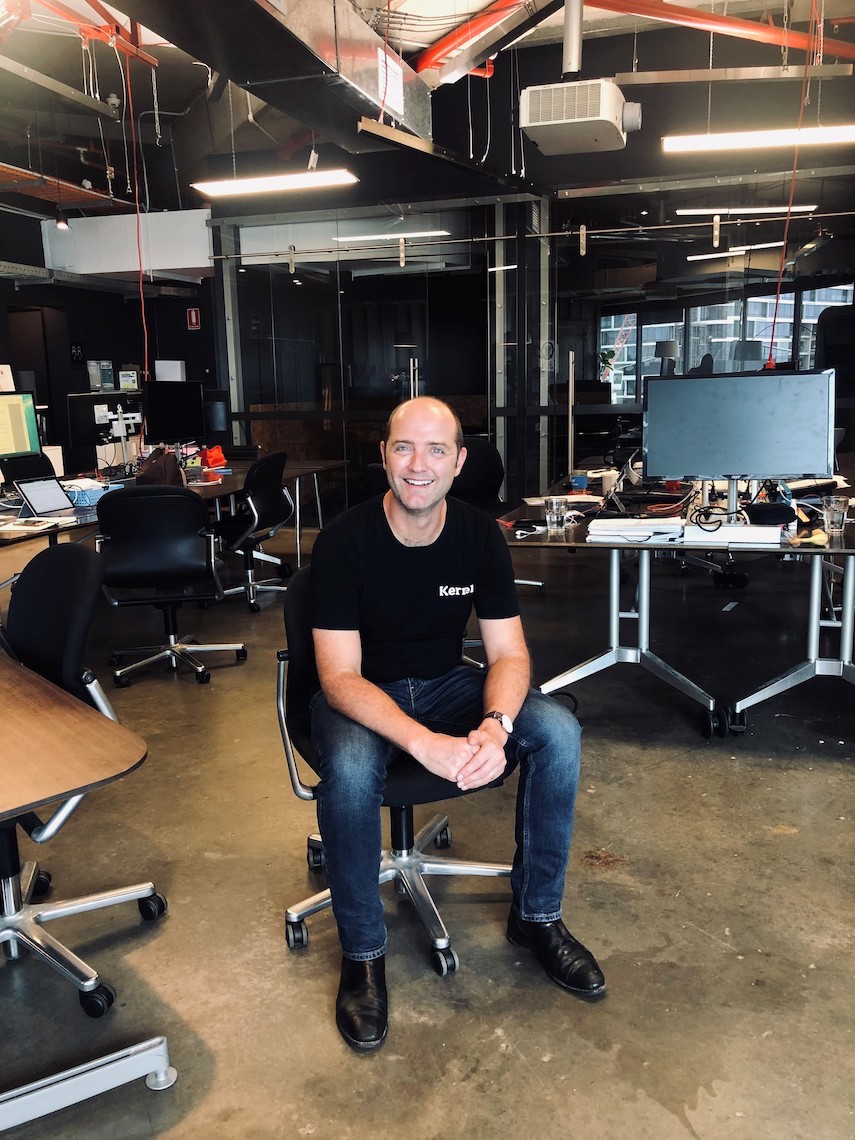 A man is sitting in an office with dark walls and many desks, in an FS swivel chair. More chairs in black can be seen in the background. Pipes and cables are visible near the ceiling.