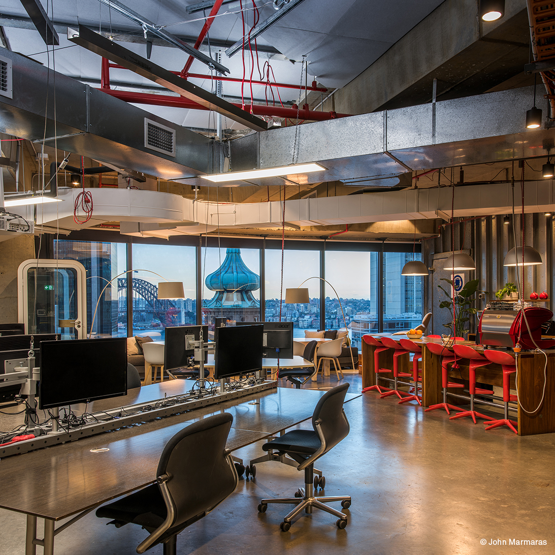 The photo shows a modern office with large windows offering a view of the city. In the foreground are several workstations equipped with black FS swivel chairs. The exposed wiring and ventilation ducts on the ceiling give the room an industrial look.
