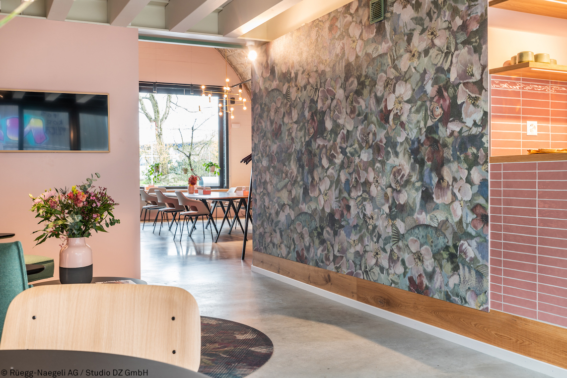 The space features a welcoming, modern entryway and lounge area with a floral accent wall, warm tones, and casual furnishings. In the foreground is an Occo chair with a wooden backrest, distinguished by its organic shell-like silhouette. The atmosphere is inviting, creative, and cozy.